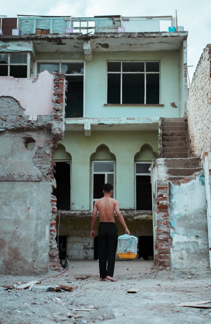A man carrying a basket in front of a decayed, abandoned two-story building.